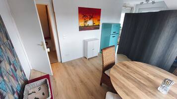 A dining area with a wooden table and chairs, a painting on the wall, and a kitchenette in the background.