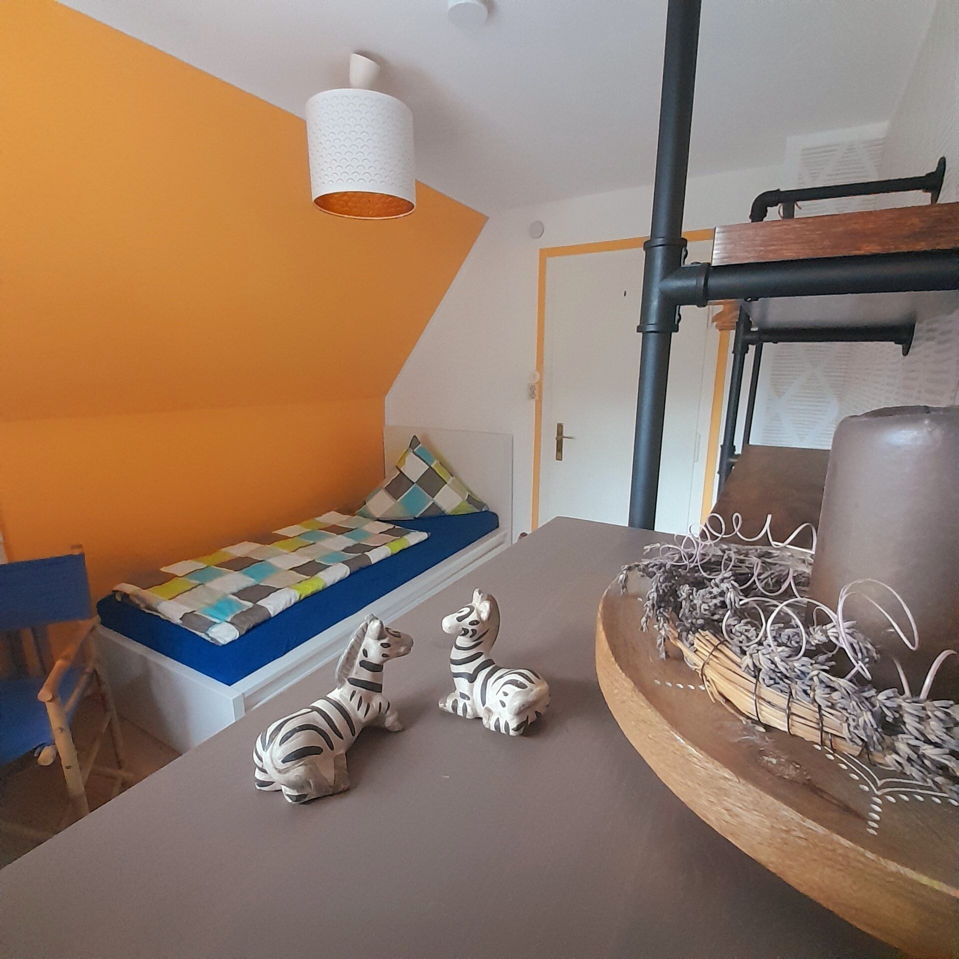 A room with a sloped ceiling, a bed with a colorful quilt, two zebra figurines, a wooden side table with a decorative item, and a white cylindrical object hanging from the ceiling.