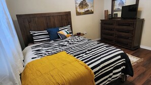 A bedroom with a bed, a wooden headboard, a ceiling fan, a nightstand, a dresser, and a painting on the wall.