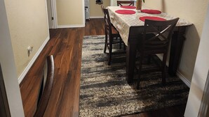 A dining area with a table set for four, a patterned rug, wooden chairs, and a painting on the wall.