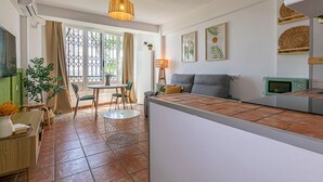 A modern living room with a grey sofa, a dining table with chairs, a small kitchen area, and a large window with curtains. - Cozy Studio in Heart of Nueva Andalucía - Pass the Keys (Marbella)