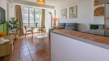 Ein modernes Wohnzimmer mit grauem Sofa, einem Esstisch mit Stühlen, einer kleinen Küchenzeile und einem großen Fenster mit Vorhängen.