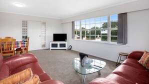 A spacious living room with a large window, a flat-screen TV, a glass coffee table, and red leather sofas. - Sunlit Glow Home with Parking and Air-con (Auckland Central Suburbs)