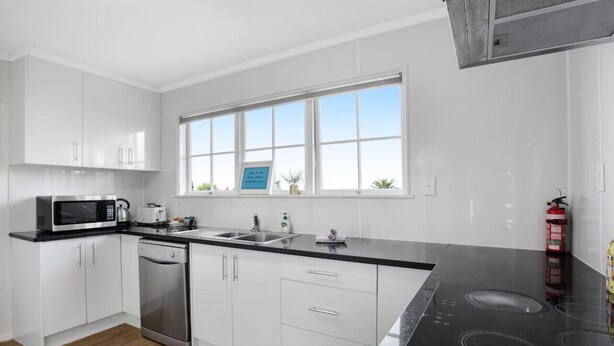 A modern kitchen with white cabinets, a black countertop, a microwave, a dishwasher, and a large window with a view. - Sunlit Glow Home with Parking and Air-con (Auckland Central Suburbs)