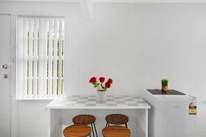 Une petite pièce propre, avec une table blanche, deux chaises, une fenêtre avec des stores, un vase de fleurs rouges et un réfrigérateur.