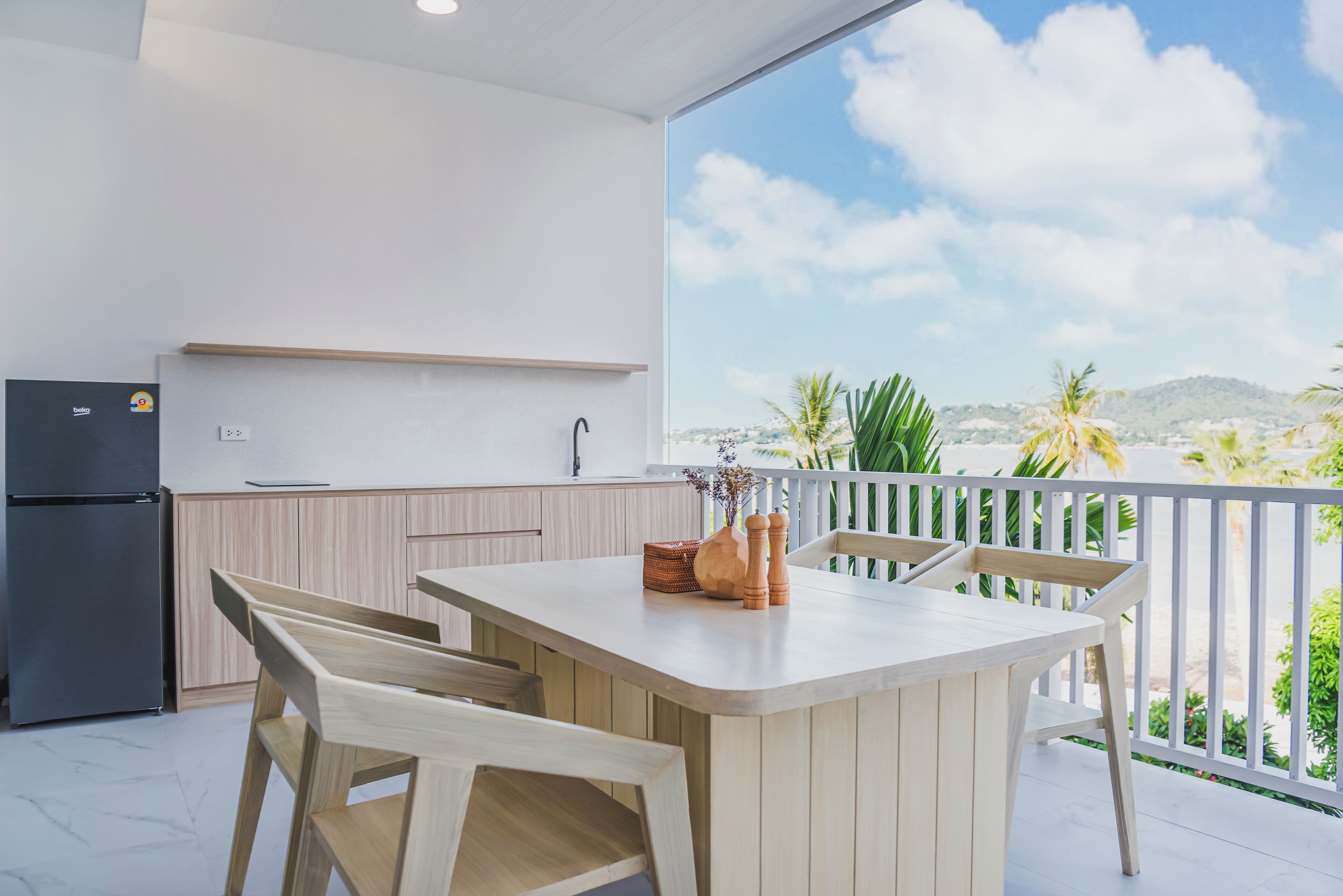 A modern kitchen with a white countertop and wooden chairs, overlooking a scenic view with palm trees and mountains.