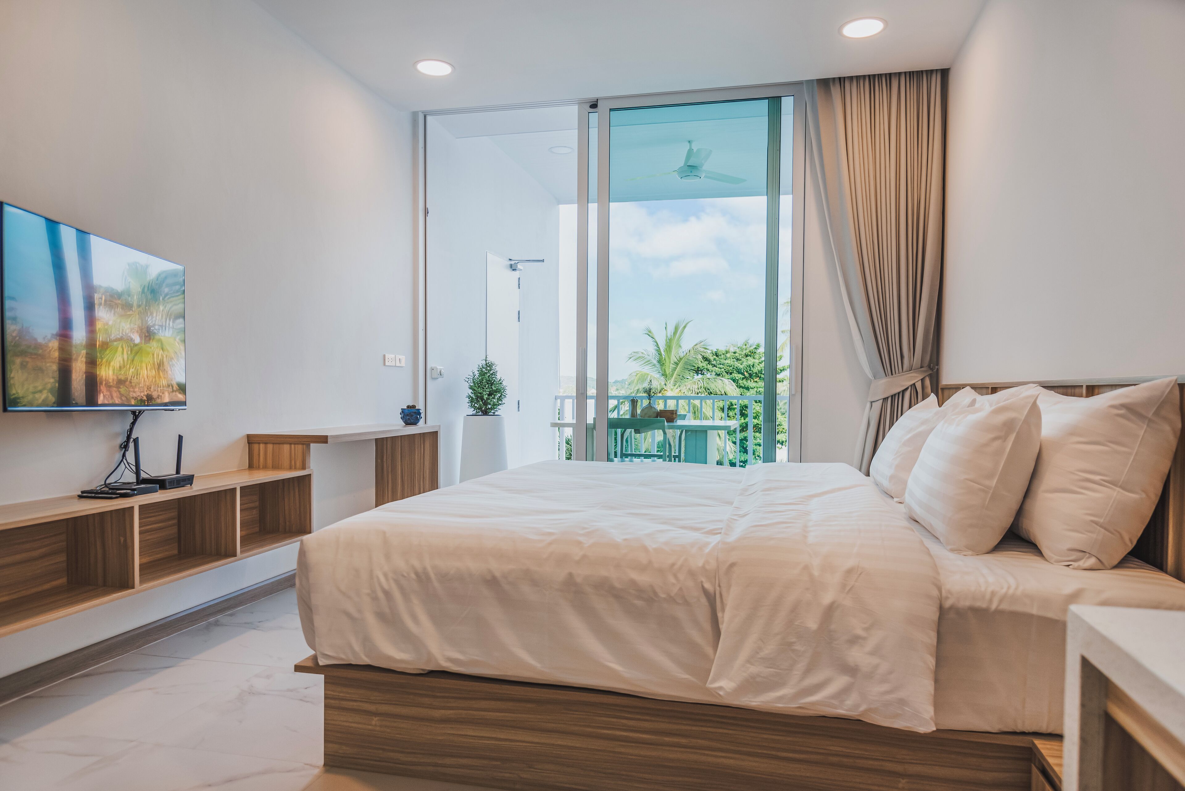 Une chambre moderne avec un grand lit, un téléviseur fixé au mur et une porte-fenêtre coulissante donnant sur un balcon avec vue sur la verdure.