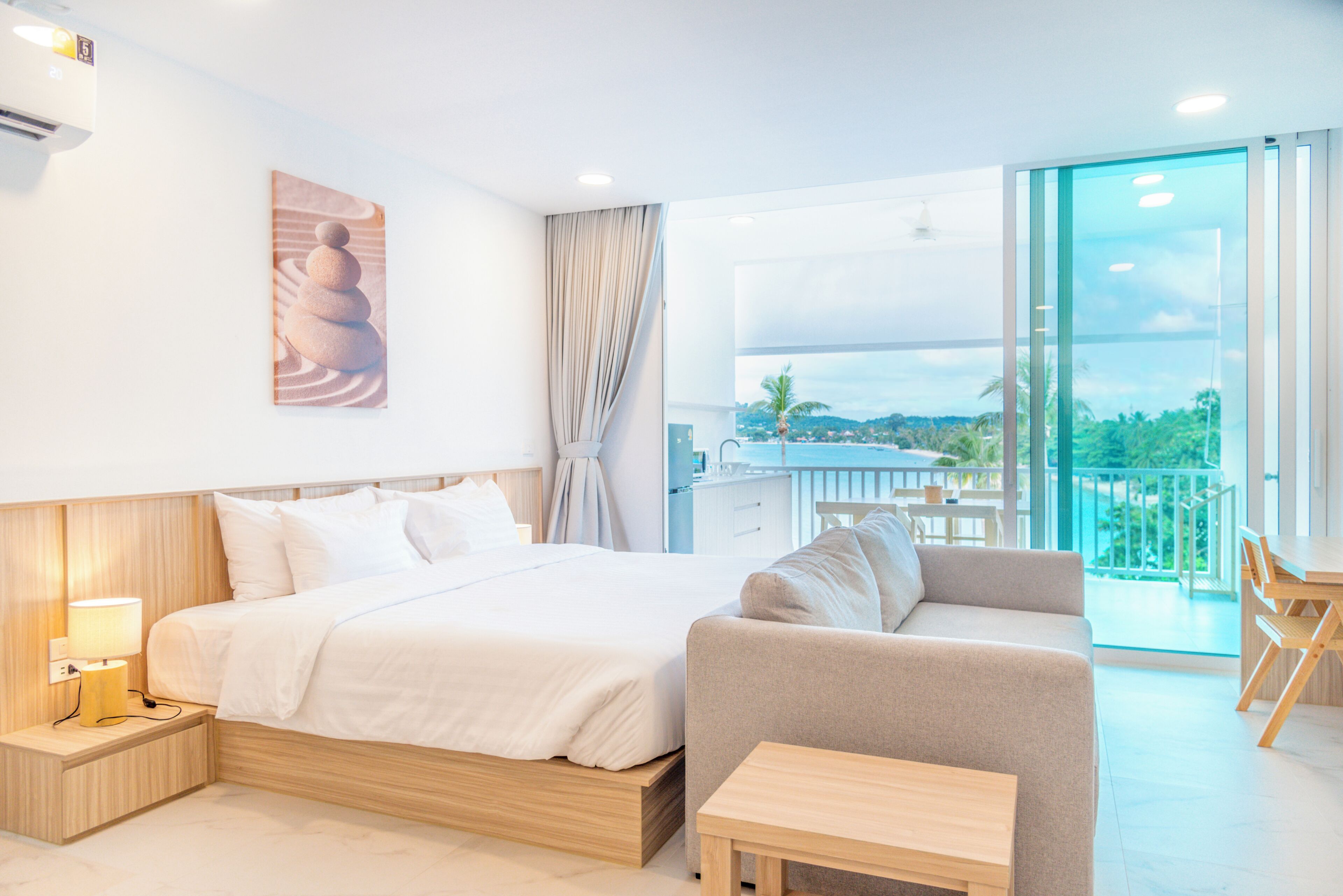 A hotel room with a bed, a sofa, a small table, a painting on the wall, and a balcony with a view.