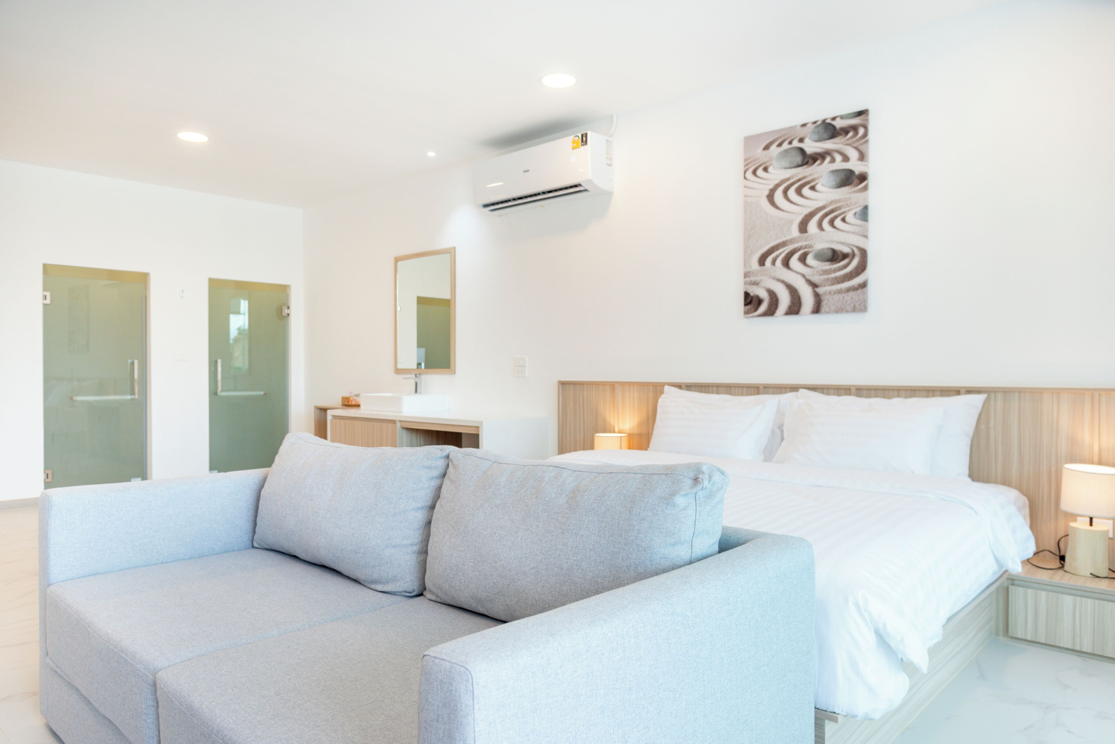 A modern hotel room with a bed, a grey sofa, a wall-mounted air conditioner, a bedside table with a lamp, and a decorative wall piece.