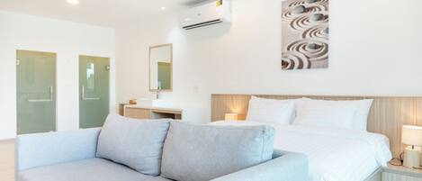A modern hotel room with a bed, a grey sofa, a wall-mounted air conditioner, a bedside table with a lamp, and a decorative wall piece.