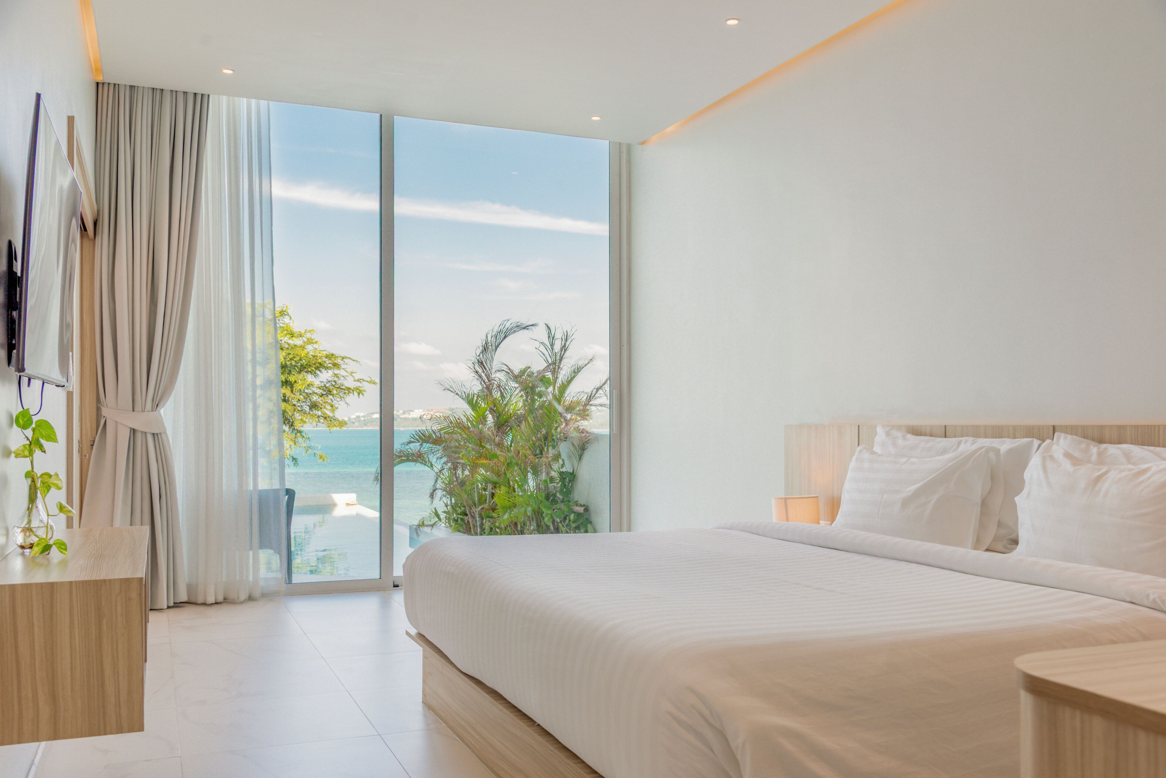 A modern hotel room with a large bed, a bedside table, a nightstand, a view of the sea, and a balcony with curtains.