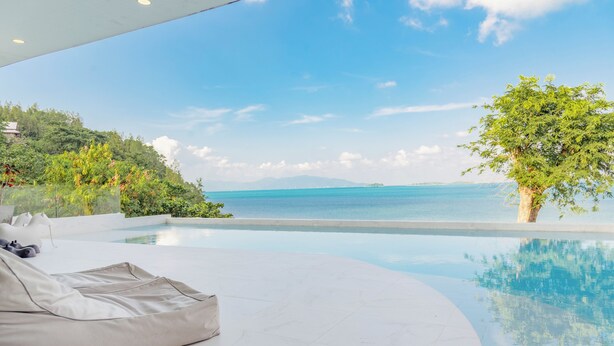 A modern pool area with a curved edge, a white lounge chair, and a view of the sea and hills. - Replay Pool Villa  (Koh Samui)