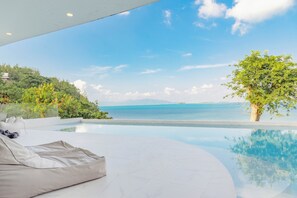 A modern pool area with a curved edge, a white lounge chair, and a view of the sea and hills. - Replay Pool Villa  (Koh Samui)