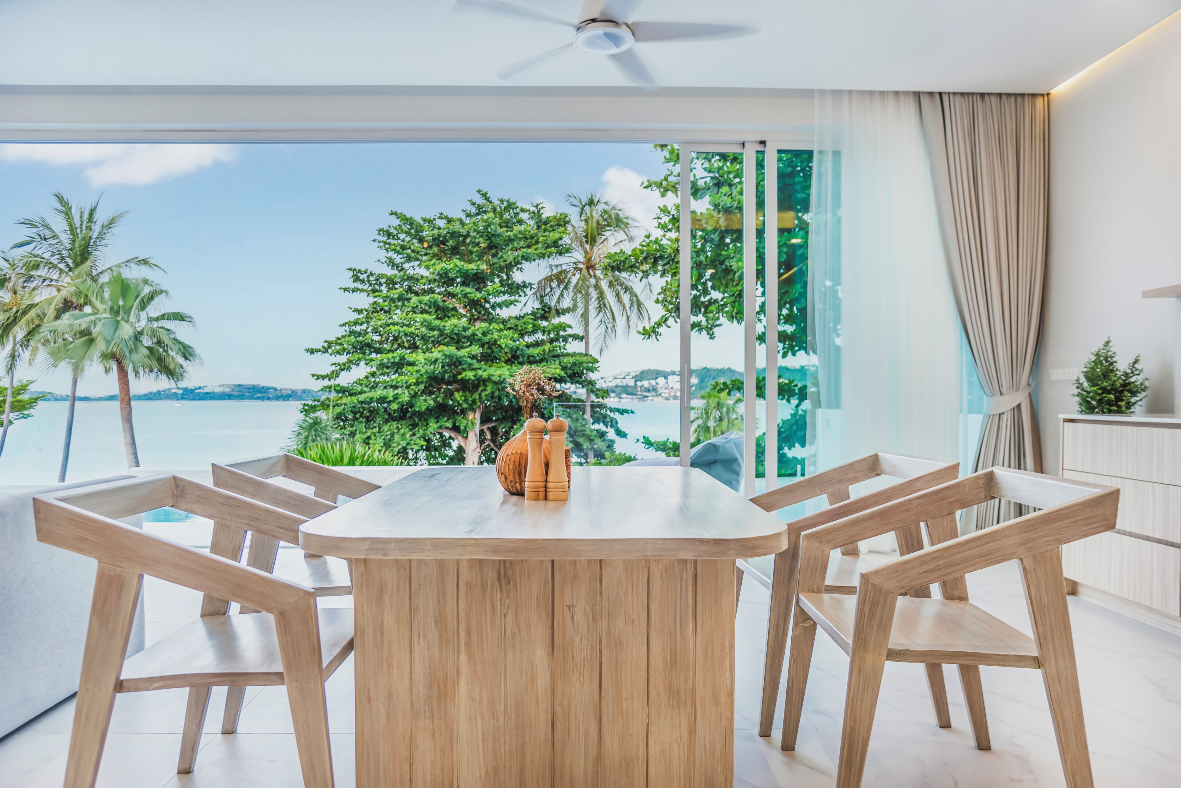 A modern dining area with a wooden table and chairs, a decorative salt and pepper set, and a view of a tropical landscape with palm trees and a body of water.