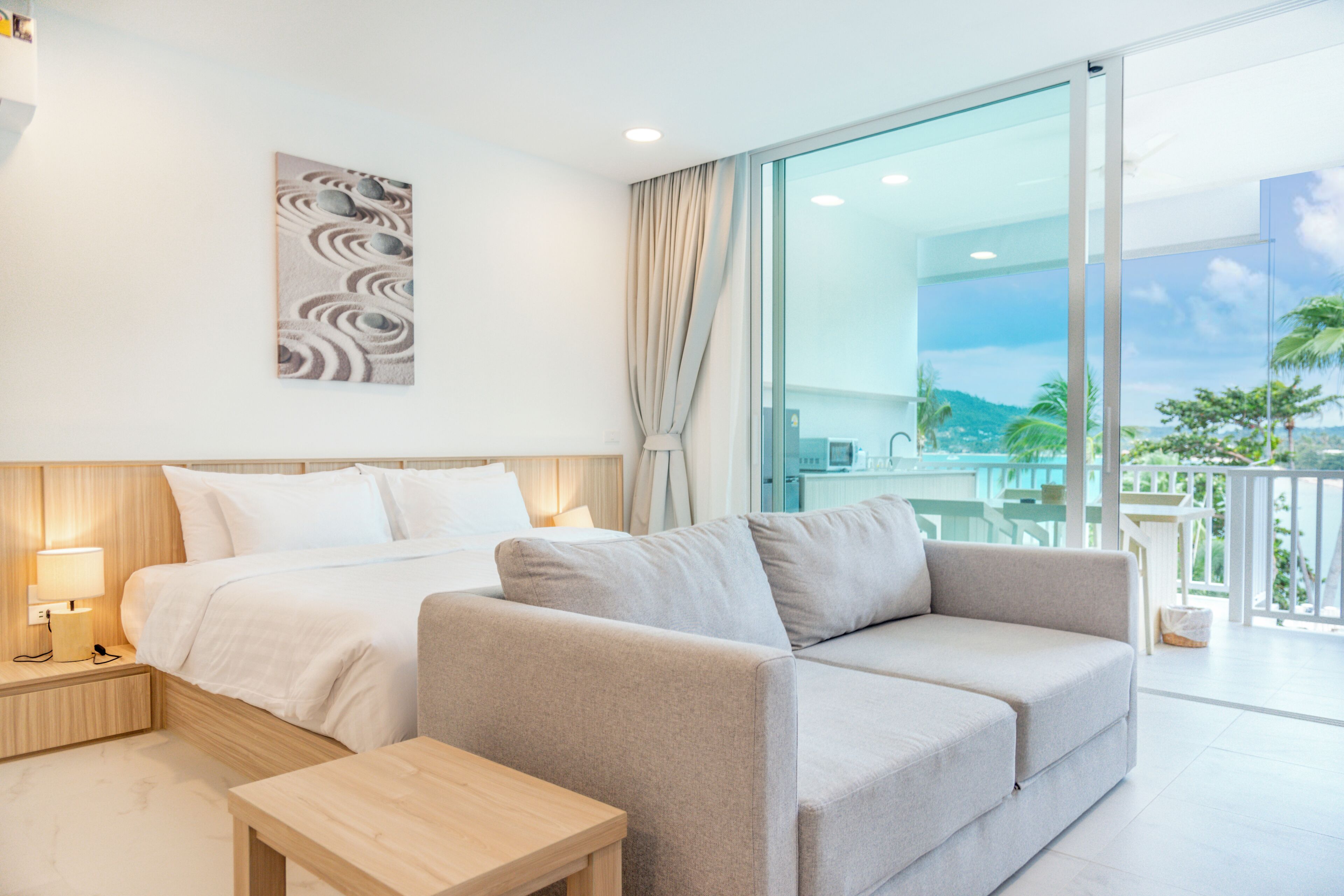A hotel room with a bed, a sofa, a small table, a nightstand, a lamp, a painting, and a large window with a view of the outdoors.