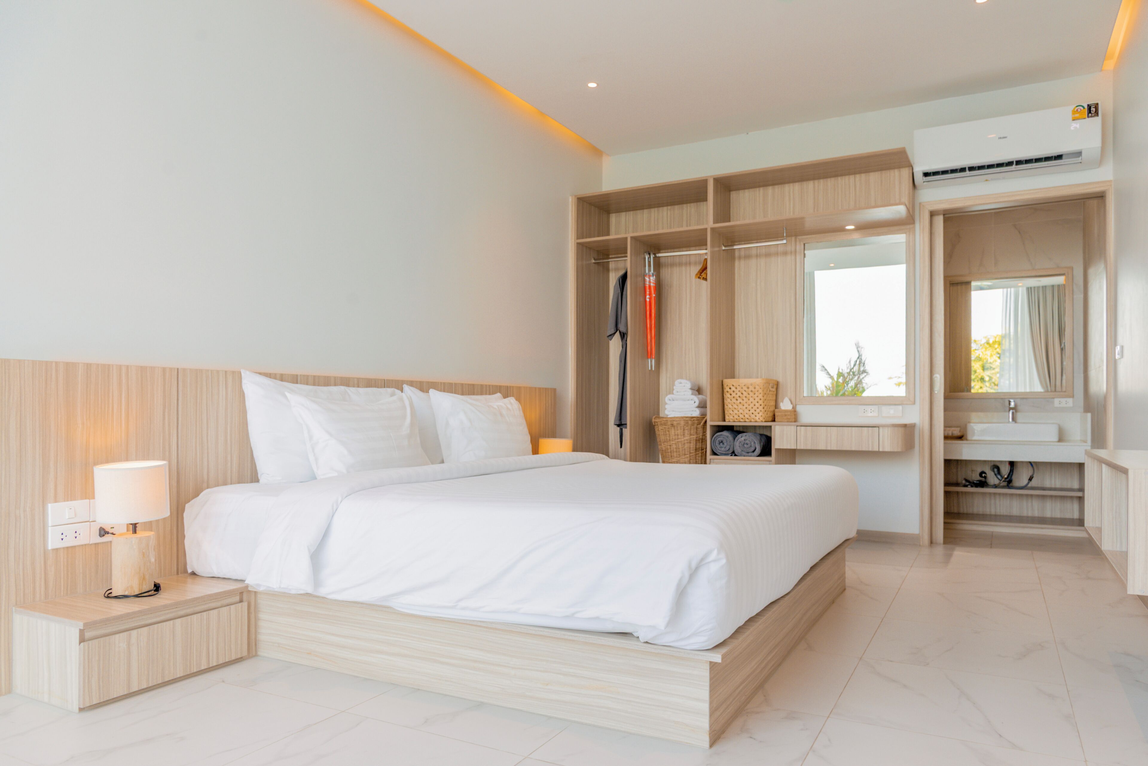 A modern hotel room with a large bed, a bedside table with a lamp, a wardrobe, and a bathroom with a mirror and sink.