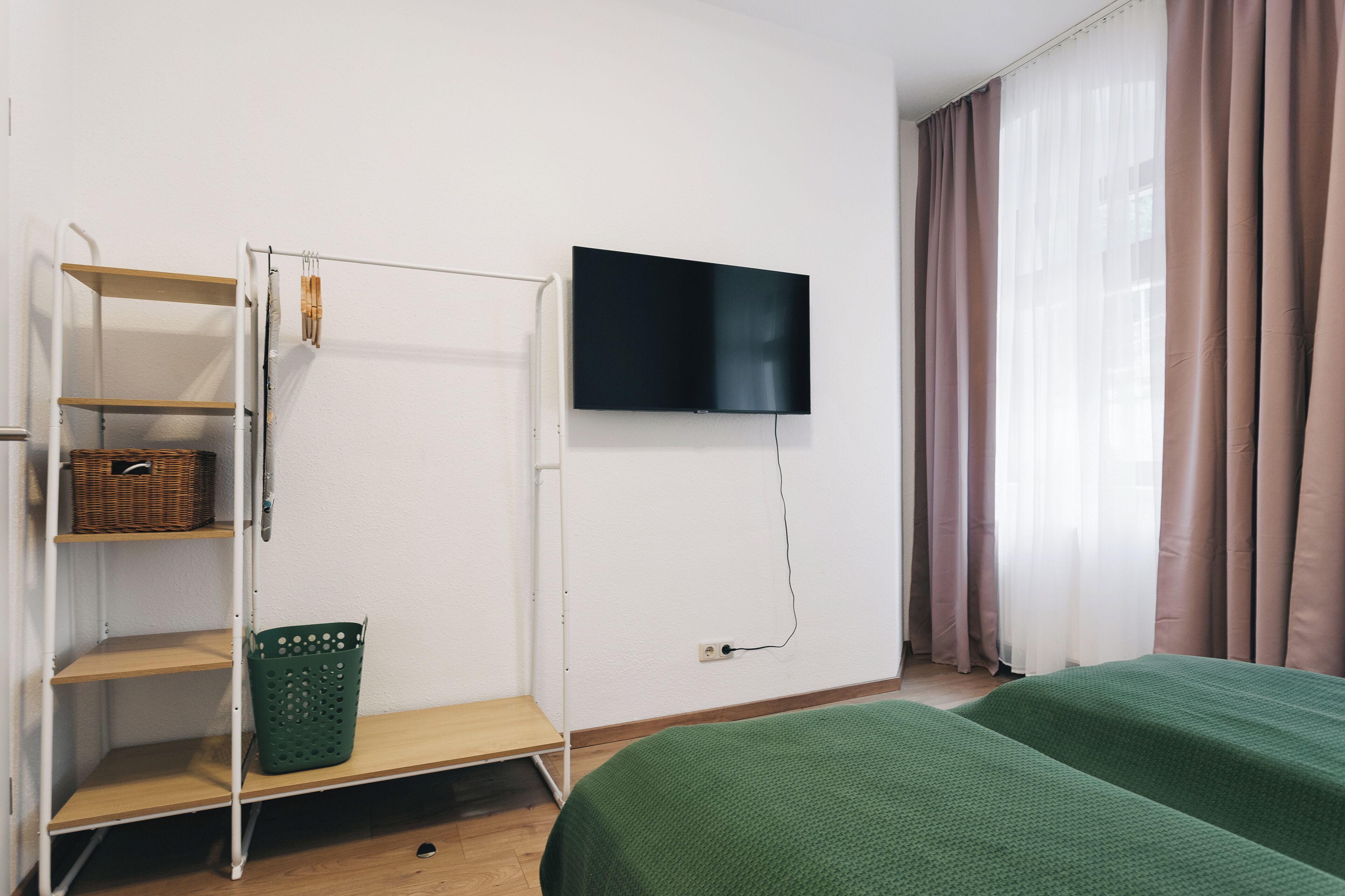 A room with a bed, a TV mounted on the wall, a shelf unit with baskets, and a window with curtains.