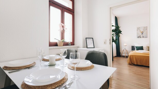 A dining area with a table set for four, a window with a plant, and a bed in the background. - Homely Apartments Neu! Wlan Küche Altstadt-sbahn-dd, Elbufer Radweg (Meißen)