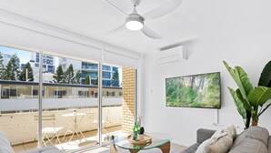 A modern living room with a glass coffee table, grey sofas, a ceiling fan, a TV, and a balcony with a view of buildings and trees. - Beachfront Escape Broadbeach - 2BD, Pool, Sleeps 7 (Gold Coast)