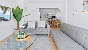 A modern living room with a grey sofa, a dining area with a wooden table, and a kitchen in the background. - Beachfront Escape Broadbeach - 2BD, Pool, Sleeps 7 (Gold Coast)