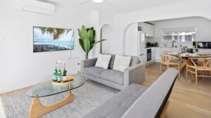 A modern living room with a grey sofa, a round coffee table, and a large plant. A kitchen with wooden cabinets and a dining area with a wooden table and chairs are visible in the background. - Beachfront Escape Broadbeach - 2BD, Pool, Sleeps 7 (Gold Coast)