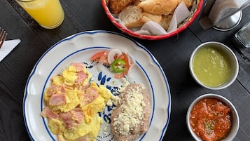 A breakfast setting with scrambled eggs, ham, and a croissant on a plate, accompanied by a glass of orange juice, a basket of bread, and two small bowls of condiments.