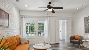 A spacious living room with a white brick wall, a ceiling fan, a large window with curtains, a grey armchair, and a round coffee table. - "Whispering Oaks"  Enjoy this 4-bedroom home with HUGE backyard (Covington)