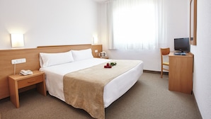 Ein Hotelzimmer mit Bett, Nachttisch mit Telefon, Schreibtisch mit Fernseher und Fenster mit Vorhängen.
