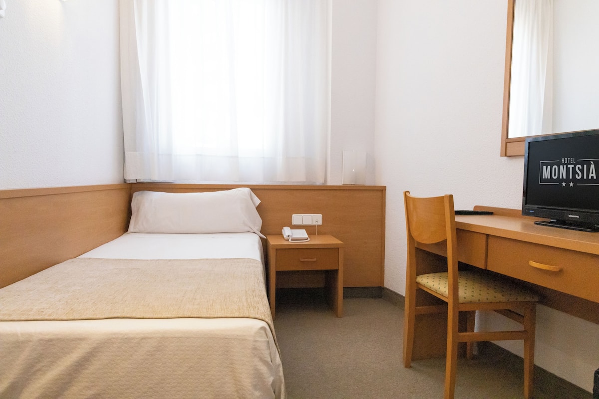 Ένα δωμάτιο ξενοδοχείου με ένα κρεβάτι, ένα γραφείο με έναν υπολογιστή, μια καρέκλα και ένα κομοδίνο με ένα τηλέφωνο.