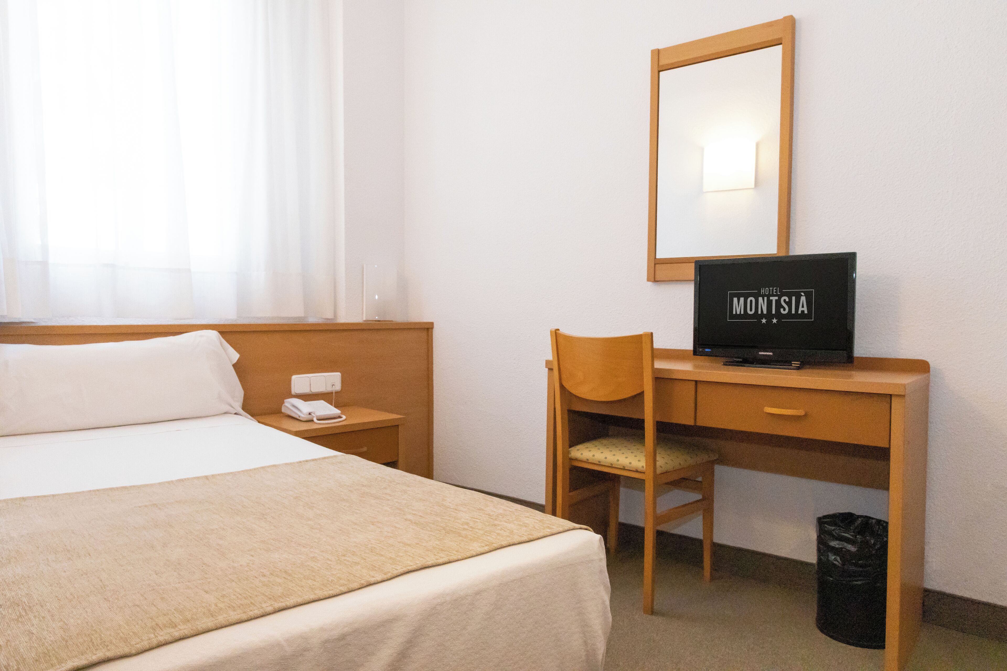 A hotel room with a bed, a desk with a computer, a chair, a mirror, and a trash bin.