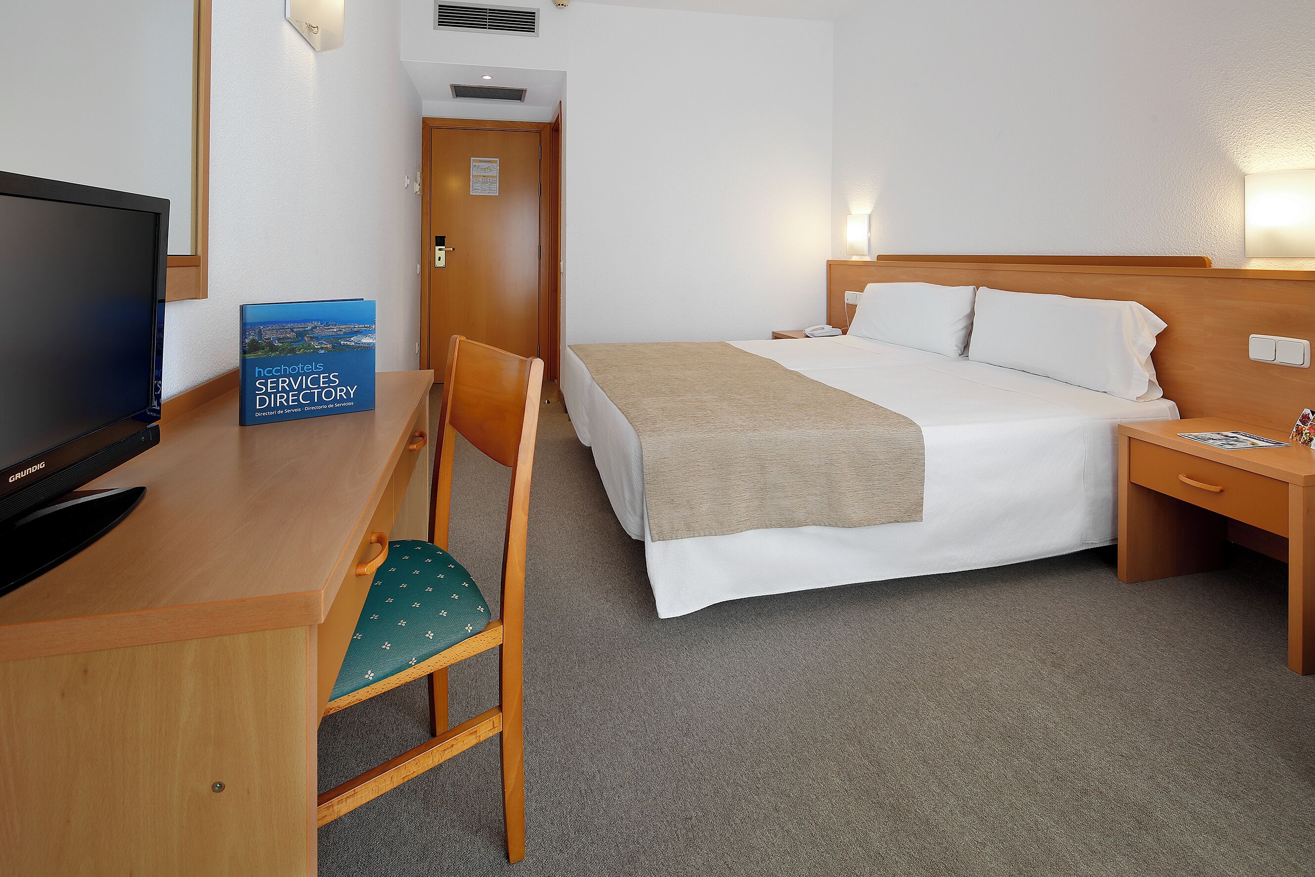 Habitación de hotel con cama, mesita de noche, silla, escritorio con televisor y un directorio sobre el escritorio.