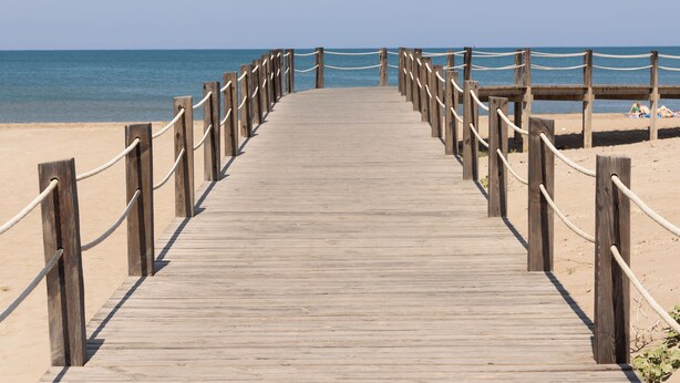 Ein Holzsteg führt zu einem Strand unter blauem Himmel.