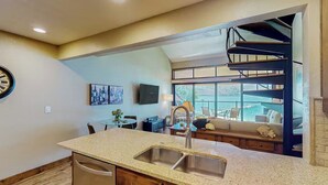 A kitchen with a granite countertop, stainless steel sink, and wooden cabinets. There is a dining area with a table and chairs, and a staircase leading to an upper level. - Lake Chelan Shores: Sandy Beach Delight # 1-5; Chelan, WA. Beautiful 2-bdr condo (Chelan)
