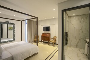 A hotel room with a large bed, a wooden dresser, two chairs, a television, and a glass-enclosed shower area.