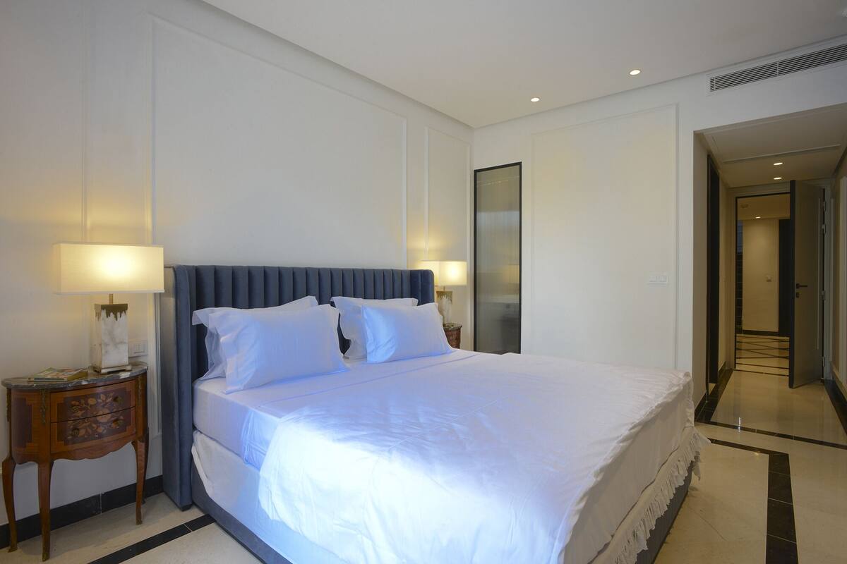 Ein Hotelzimmer mit einem großen Bett, zwei Nachttischen mit Lampen, einem Spiegel und einer Tür zu einem anderen Raum.