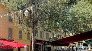 Ein Straßencafé mit Tischen und Stühlen, ein rotes Sonnensegel und ein Baum im Hintergrund.