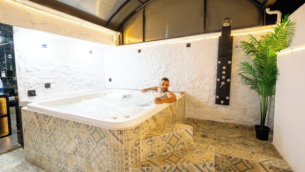 A man relaxing in a large, freestanding hot tub with patterned tiles, set against a white textured wall and a potted plant. - HOTEL LAX  (Medellín)