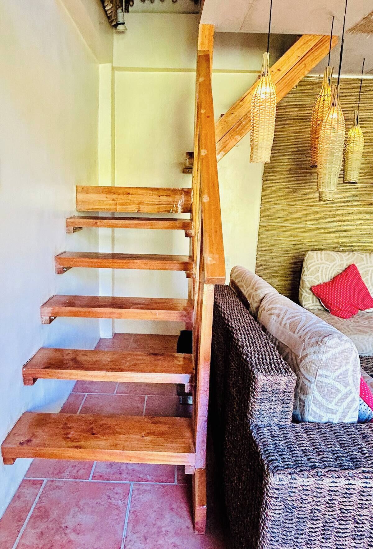 Un escalier en bois, doté d’une rampe, mène à un espace de vie comprenant un canapé et des coussins.