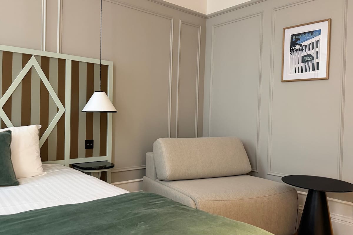 Een hotelkamer met een bed, een stoel, een nachtkastje, een lamp en een ingelijst schilderij aan de muur.