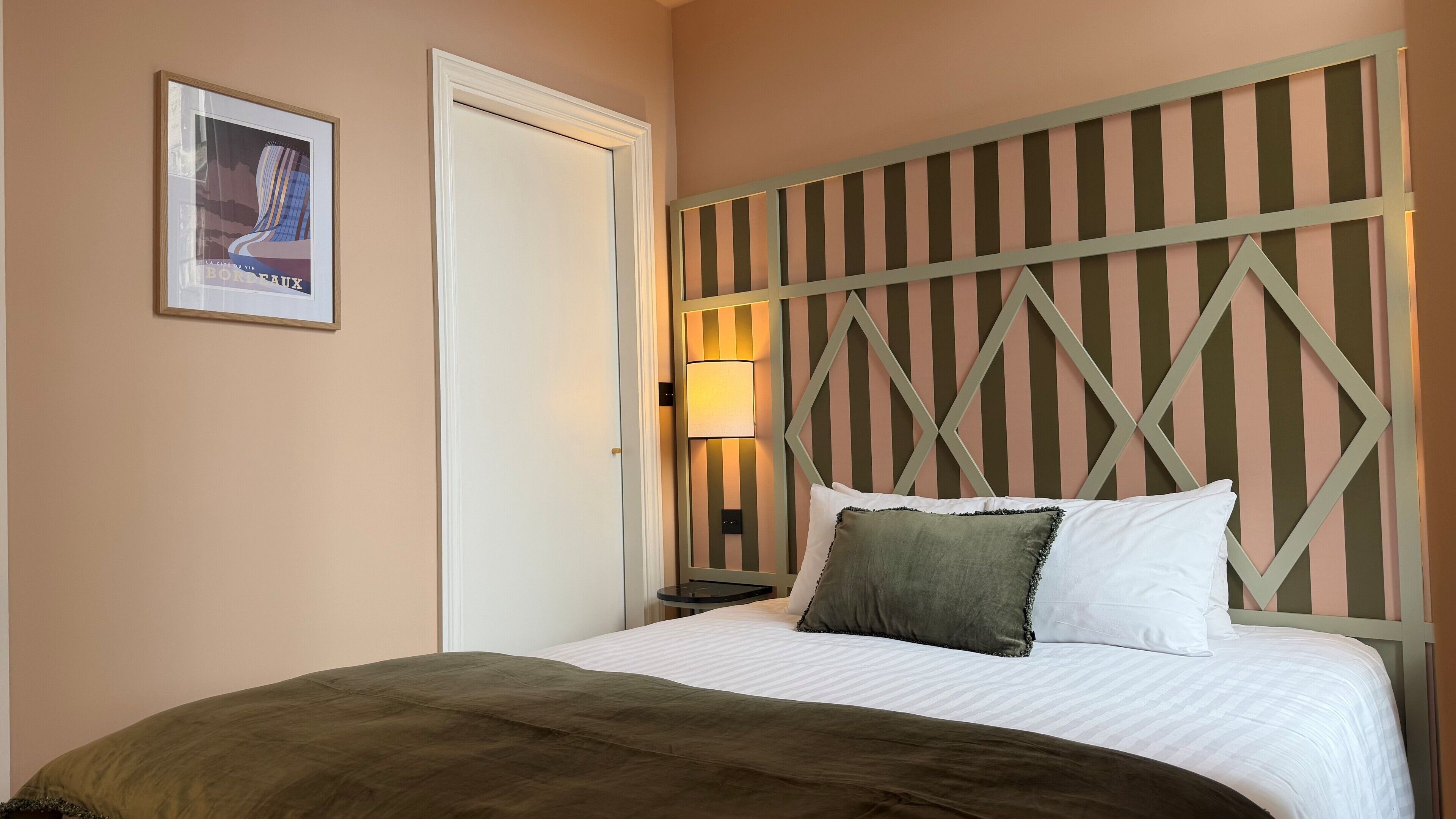 Quarto de hotel com cama, mesa de cabeceira, abajur e quadro na parede.