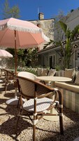 Terraza al aire libre con sillas de mimbre y un gran paraguas rosa, rodeada de follaje y un patio empedrado.