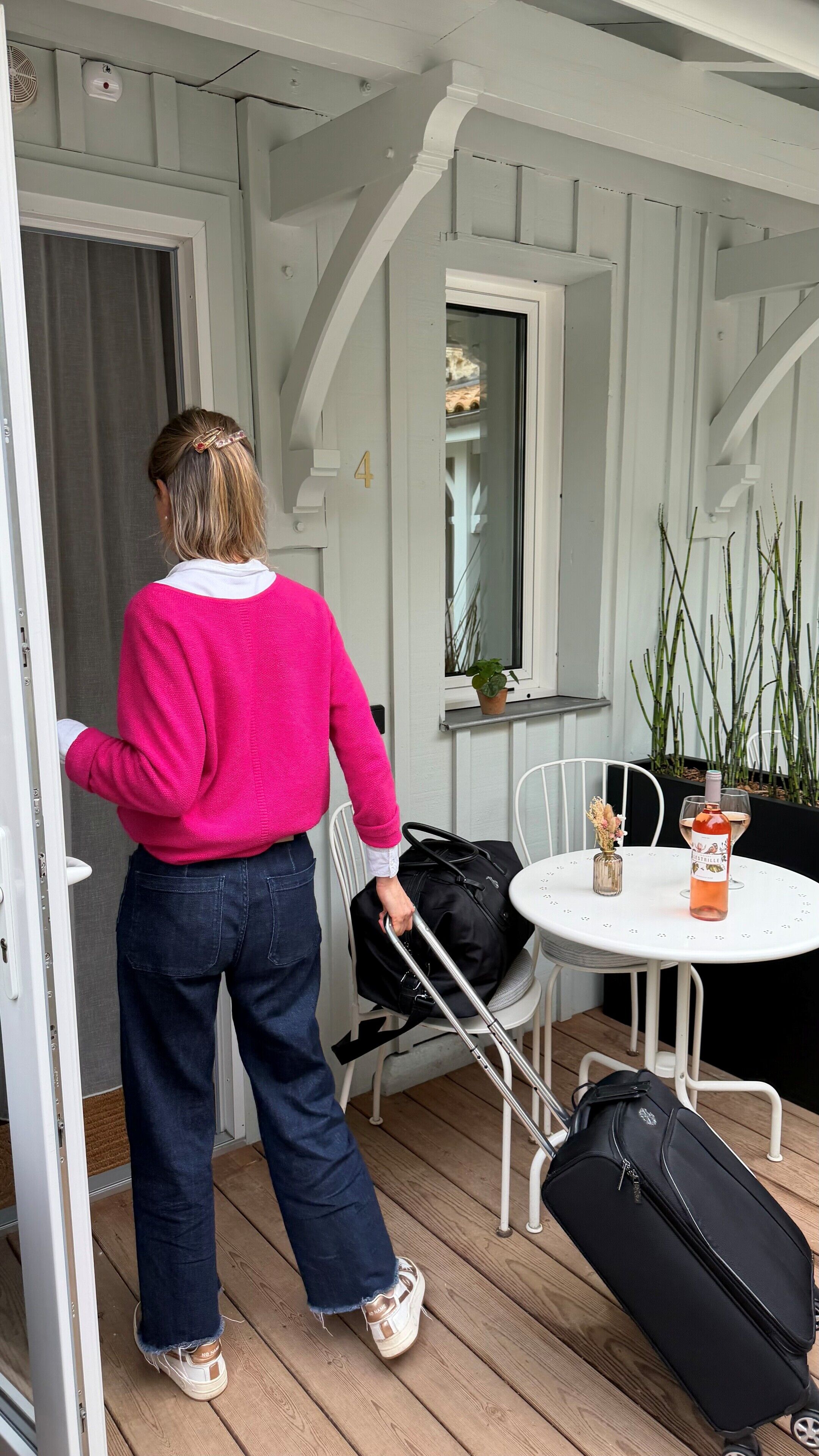 Une personne sort d’une pièce, une valise à la main. On aperçoit une table avec une bouteille et un verre, ainsi qu’une fenêtre avec une plante.