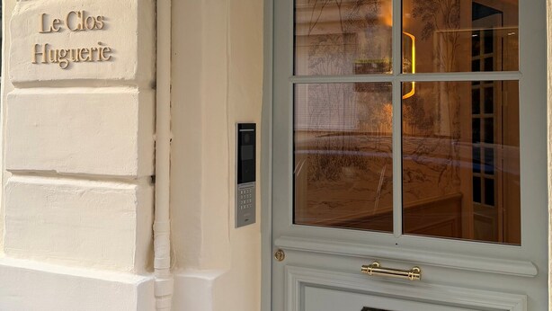 Een witte deur met een ruit en een messing handgreep, een wandlamp en een bordje met de tekst 'Le Clos Huguerie'.