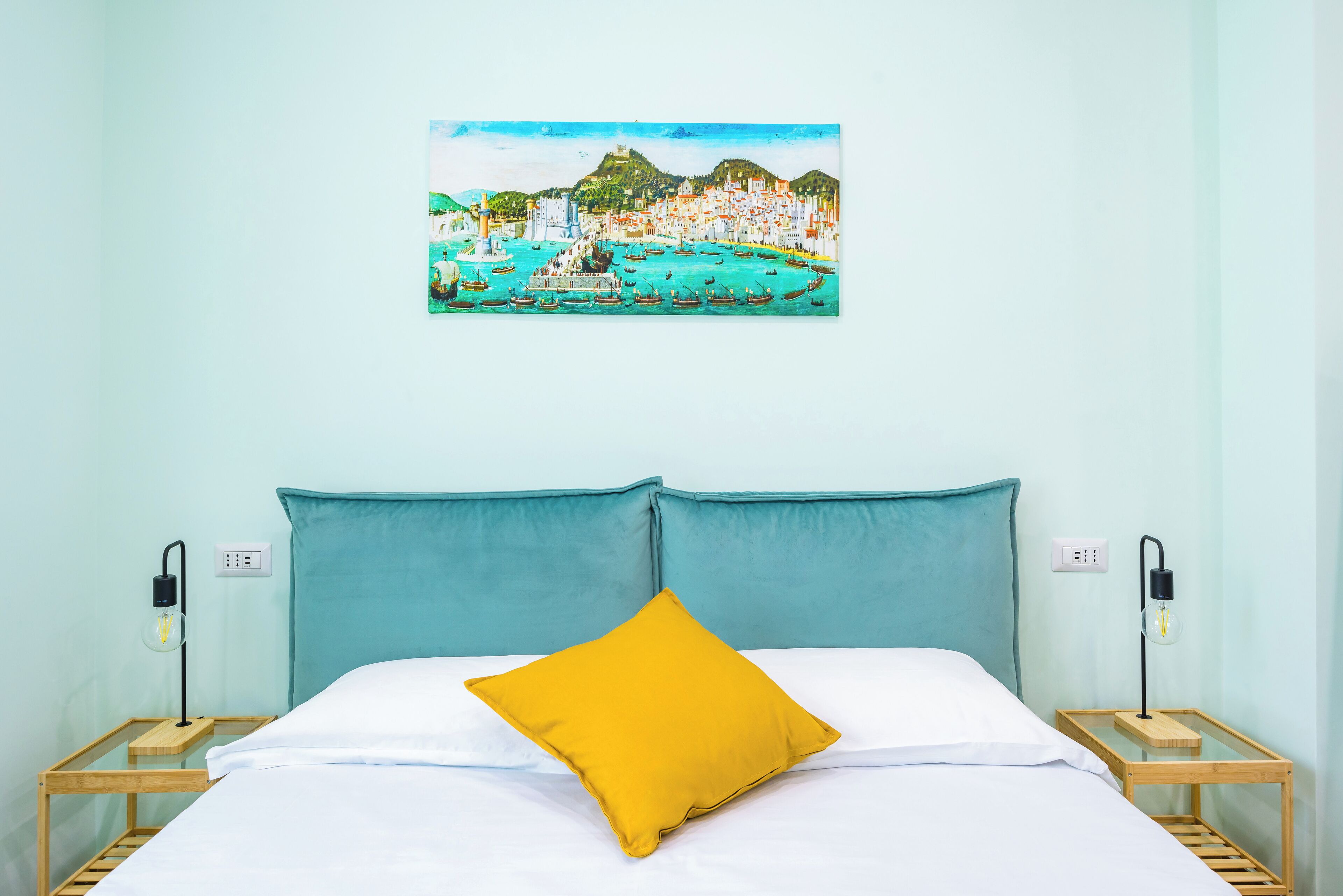 Une chambre à coucher avec un lit, une tête de lit de couleur turquoise, un oreiller jaune et un tableau représentant une ville côtière accroché au mur.