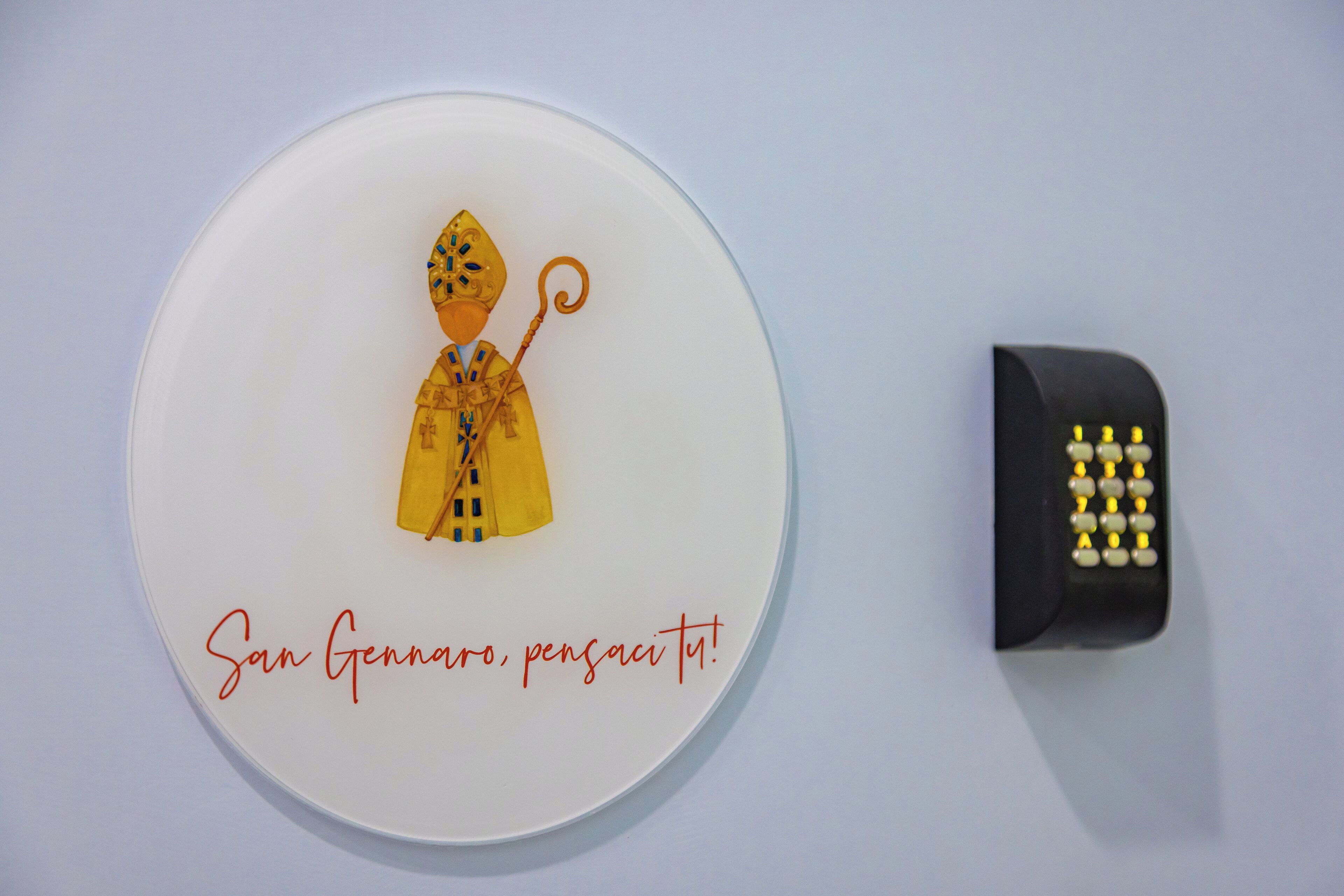A wall-mounted artwork of a figure holding an umbrella, accompanied by the text "San Giovanni, pensaci tu!" and a remote control.