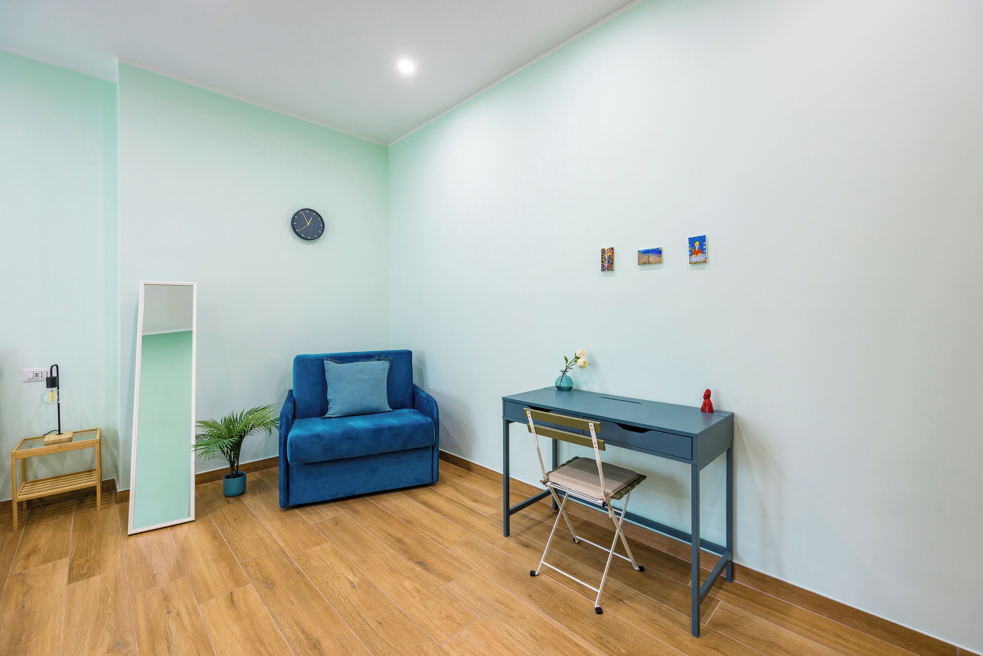 A room with a blue sofa, a desk with a chair, a small plant, and a clock on the wall.