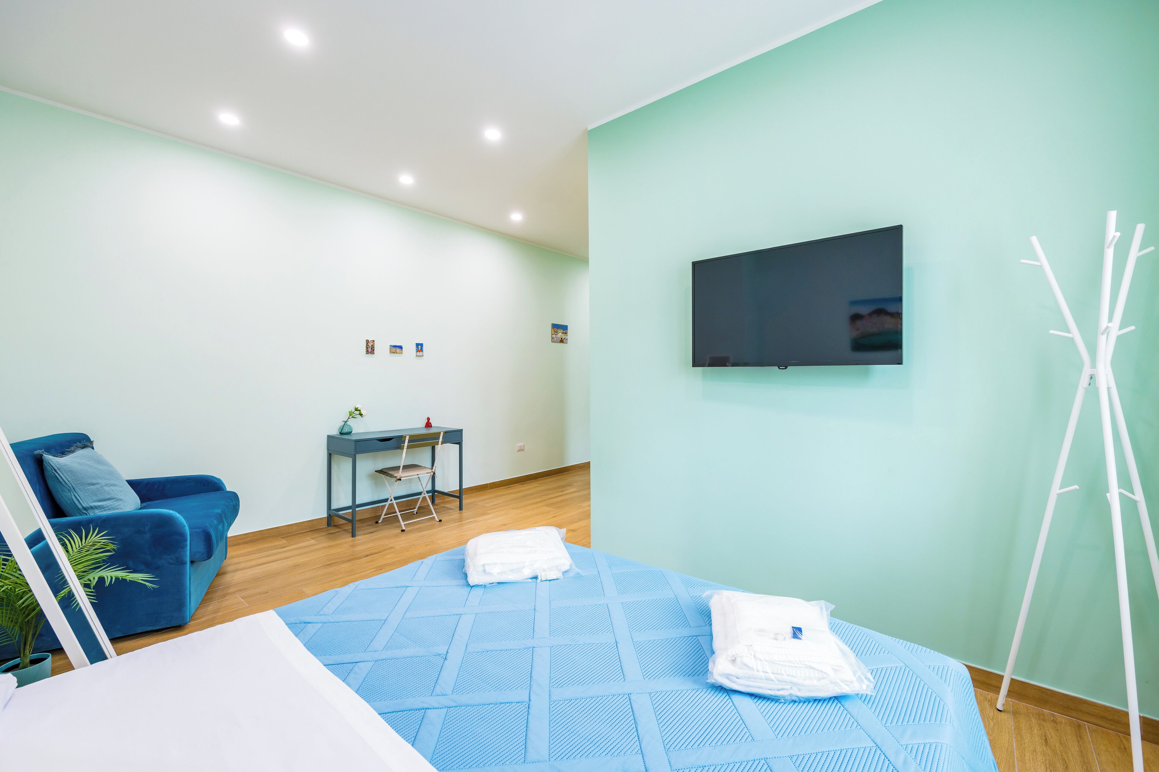 Une chambre d’hôtel moderne avec un lit bleu, un canapé bleu, un bureau avec une chaise, un téléviseur fixé au mur et une plante blanche.