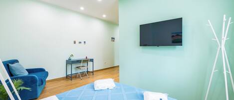 Una habitación de hotel moderna con una cama y un sofá azules, un escritorio con una silla, un televisor en la pared y una planta blanca.