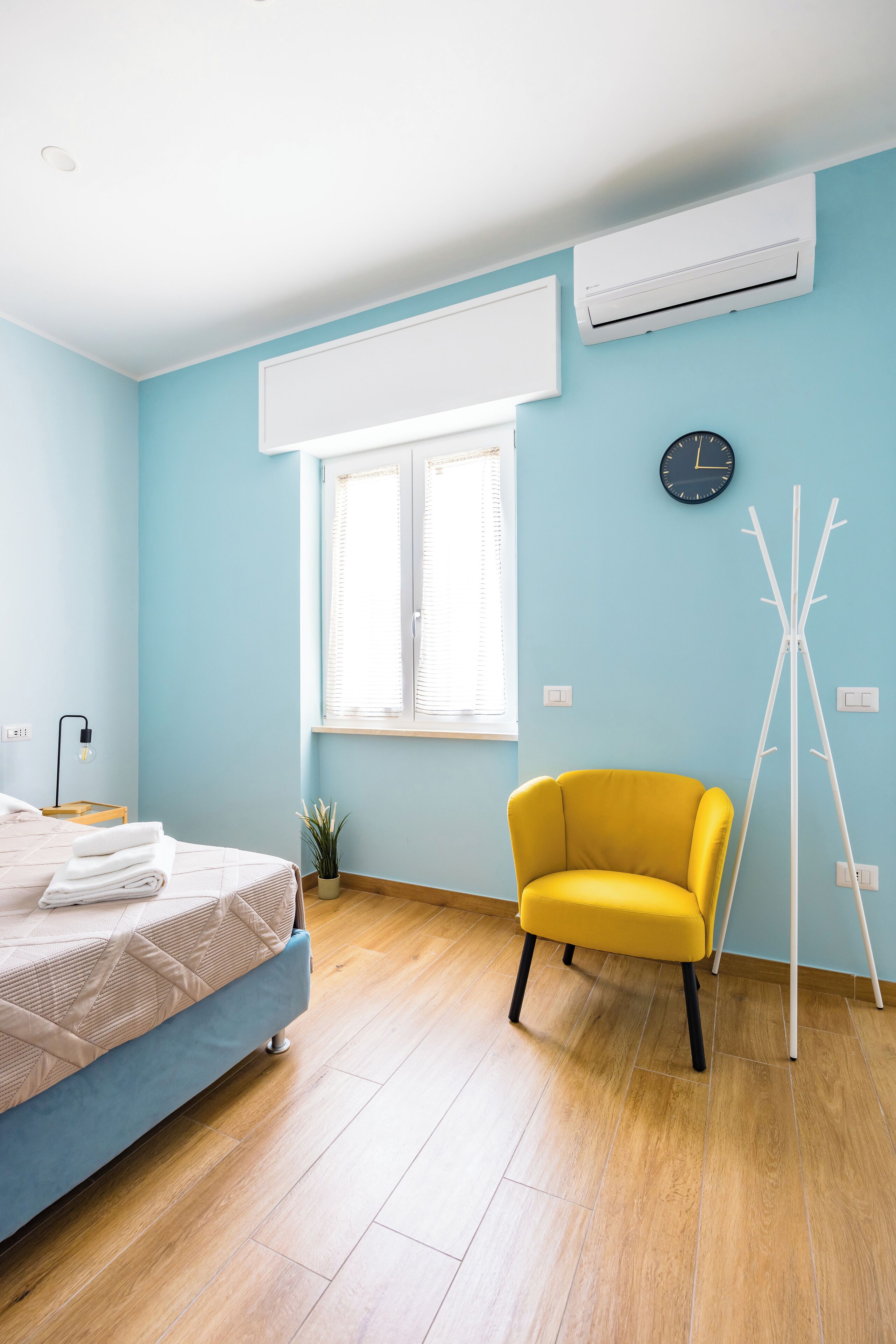 A room with a bed, a yellow chair, a window with blinds, an air conditioning unit, and a clock on the wall.