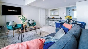 Een moderne woonkamer met een bank, stoelen en een salontafel. Er is een eethoek met een tafel en stoelen. Een televisie is aan de muur bevestigd.